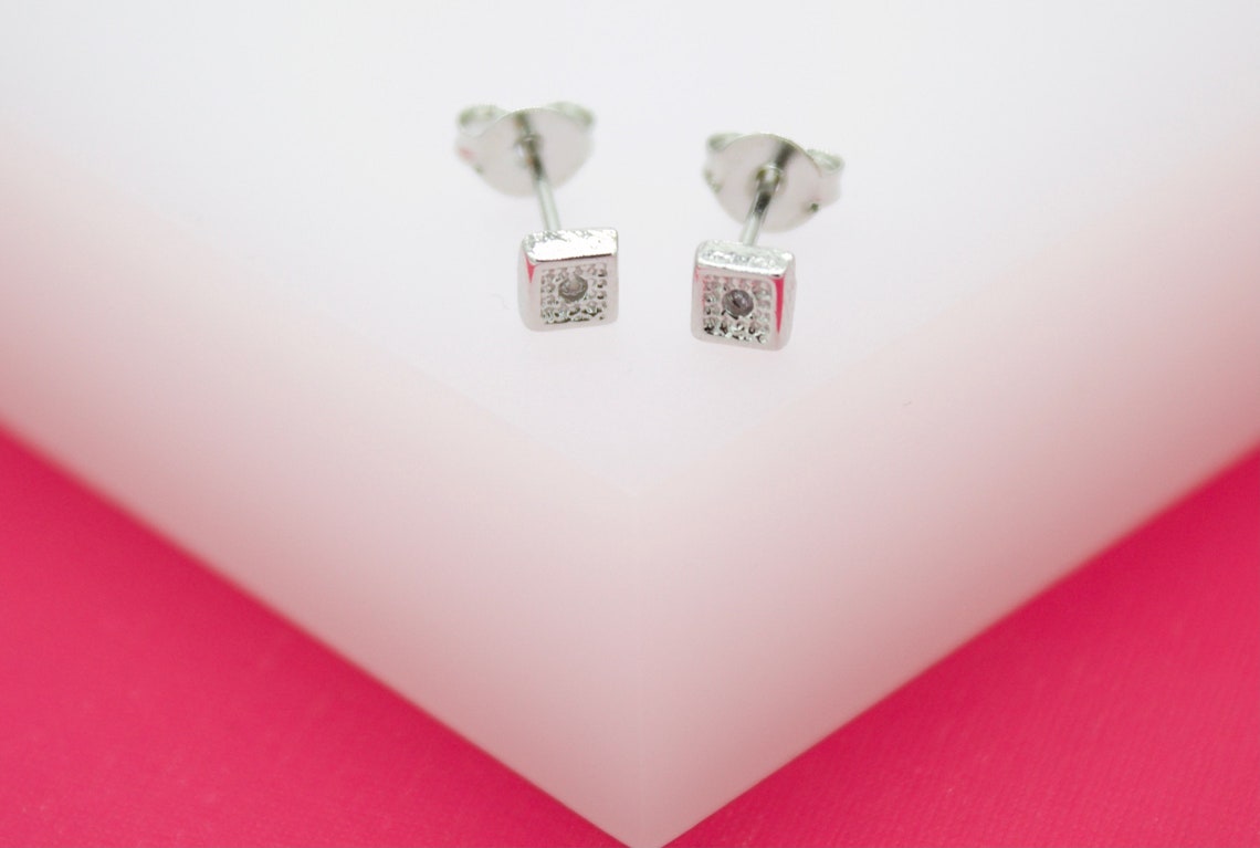 A pair of delicate silver square stud earrings rests on a white and pink display surface.