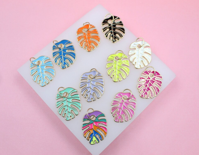 A collection of colorful enamel monstera leaf charms with gold trim rests on a white display block against a pink background.