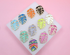 A collection of colorful enamel monstera leaf charms with gold trim rests on a white display block against a pink background.