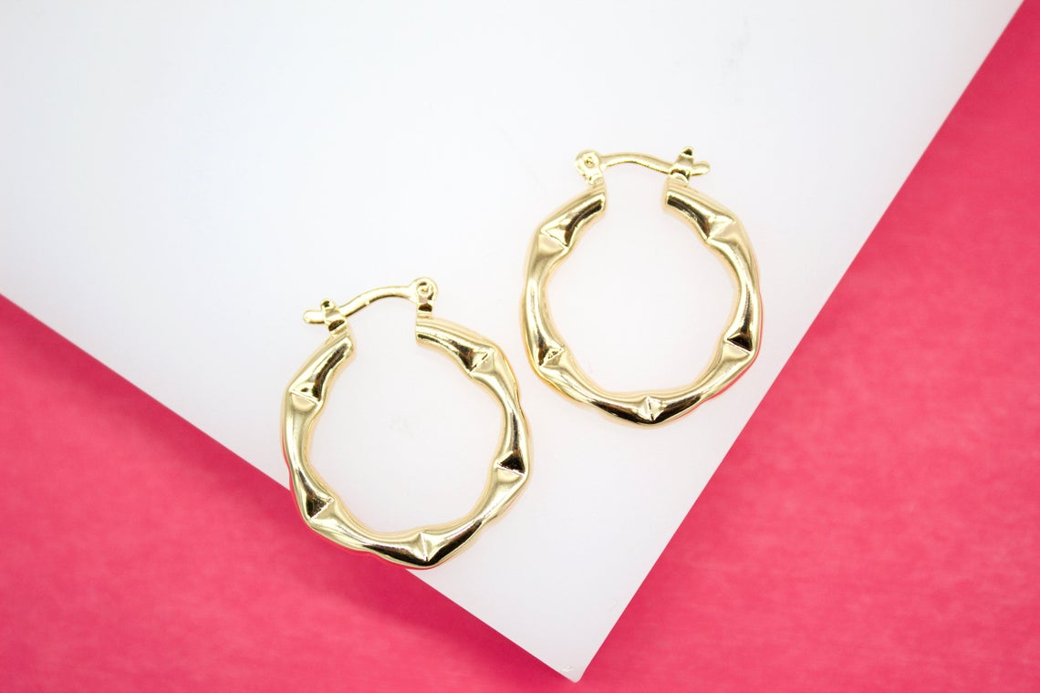 Two polished gold bamboo hoop earrings rest on a white display surface against a vibrant pink background.