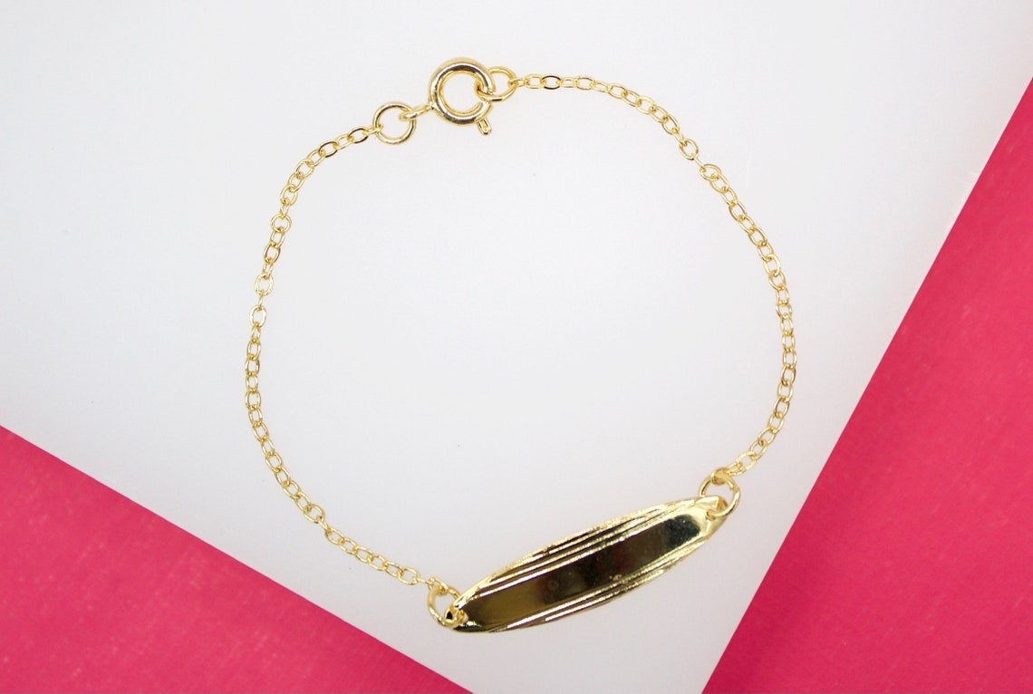 A delicate gold ID bracelet rests on a white and pink surface, showcasing its polished metal plate and fine chain link.