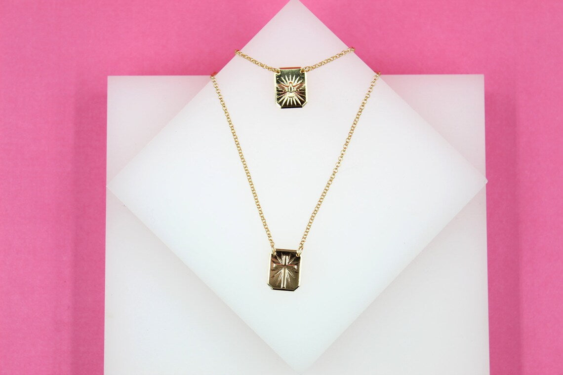 A layered gold necklace featuring rectangular pendant charms with sunburst engravings rests on a white display against a vibrant pink background.