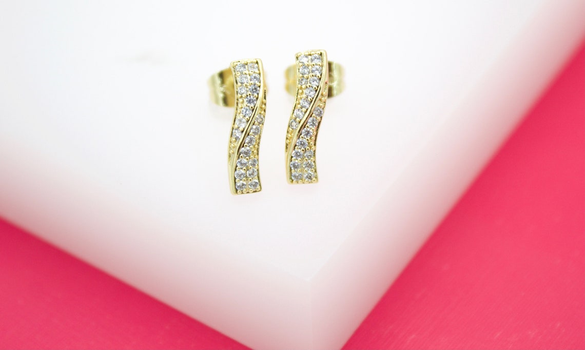 A pair of elegant gold wave stud earrings embellished with sparkling cubic zirconia stones rests on a white and pink display stand.