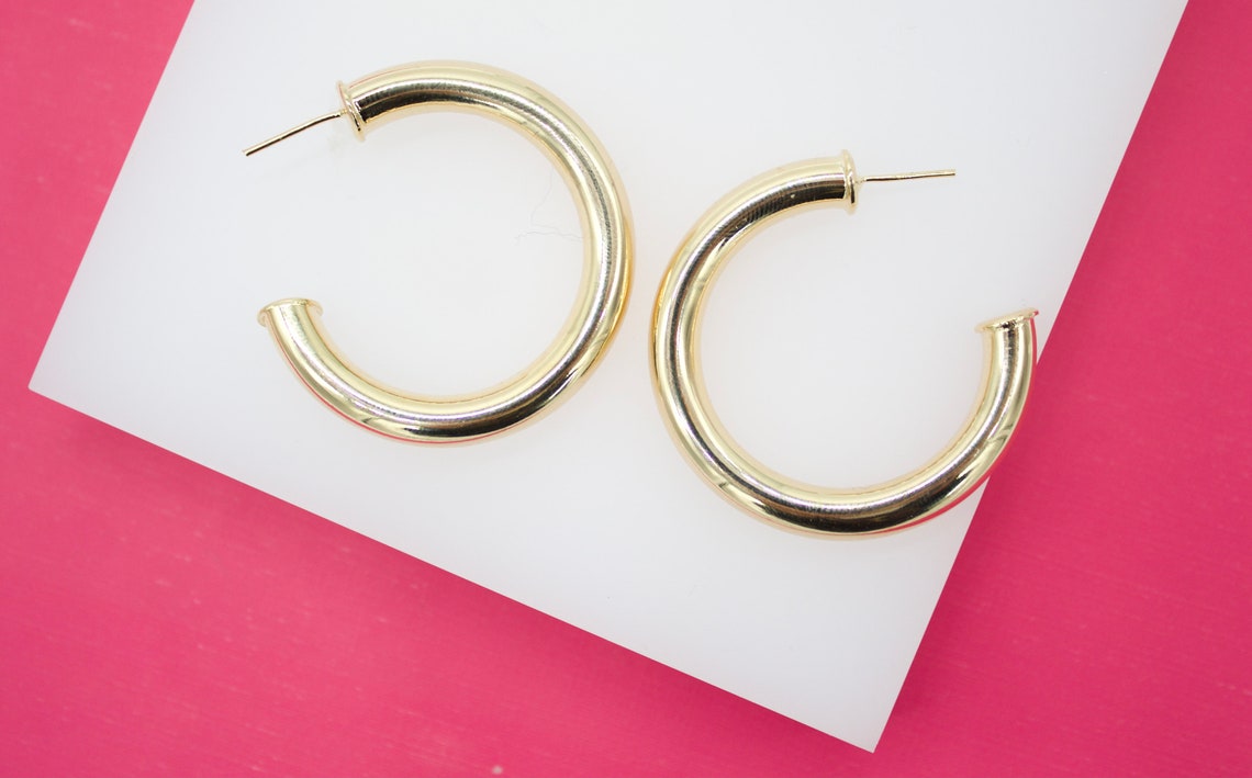 A pair of chunky gold hoop earrings rests against a white display block on a vibrant pink background.