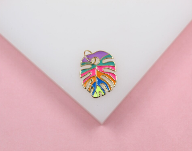 A colorful enamel monstera leaf pendant rests on a white display surface against a pink background.