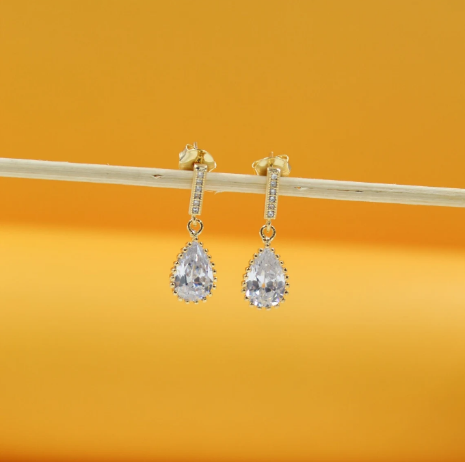 A pair of gold drop earrings featuring dangling pear-shaped cubic zirconia crystals hang from a thin wooden stick against a warm yellow background.
