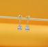 A pair of gold drop earrings featuring dangling pear-shaped cubic zirconia crystals hang from a thin wooden stick against a warm yellow background.