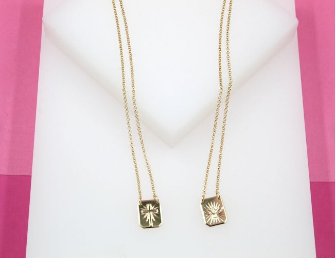 Two delicate gold necklaces featuring engraved rectangular pendants displaying a cross and a religious motif hang against a white and pink background.