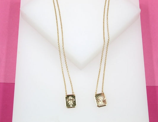 Two delicate gold necklaces featuring engraved rectangular pendants displaying a cross and a religious motif hang against a white and pink background.