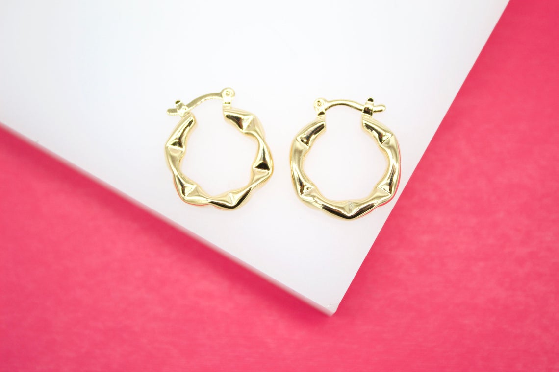 A pair of textured gold hoop earrings rests on a white display block against a vibrant pink background.