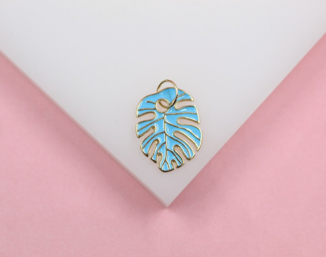 A small gold and blue enamel monstera leaf charm rests on a white display stand.