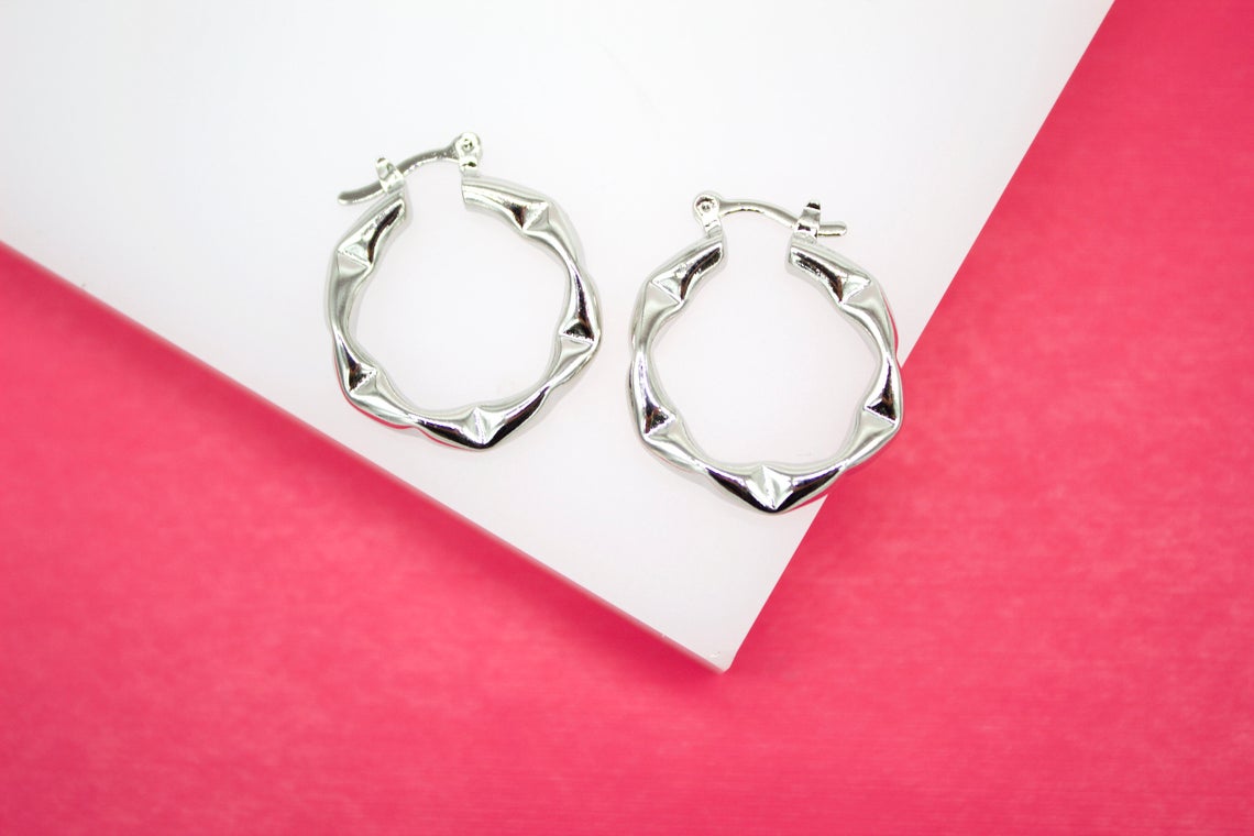 A pair of silver bamboo hoop earrings rests on a white display surface against a vibrant pink background.