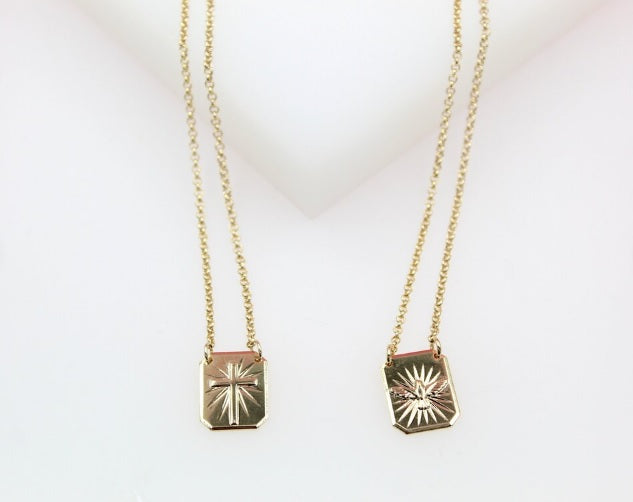 Two delicate gold charm necklaces featuring rectangular pendants with a cross and a sunburst design display against a light background.