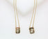 Two delicate gold charm necklaces featuring rectangular pendants with a cross and a sunburst design display against a light background.