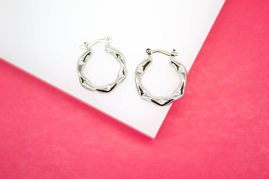 A pair of shiny silver bamboo hoop earrings rests on a white platform against a vibrant pink background.