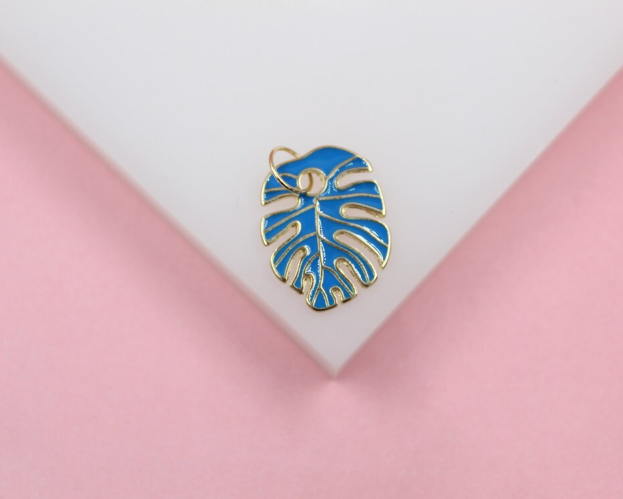 A vibrant blue and gold monstera leaf charm rests on a white and pink surface.