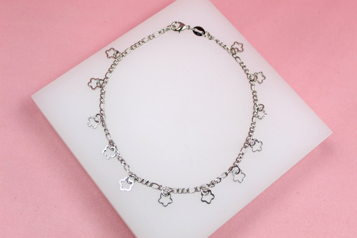 A silver charm bracelet with delicate flower-shaped charms rests on a white display stand against a pink background.