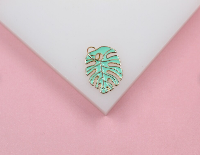 A mint green and gold monstera leaf charm rests on a white and pink display stand.