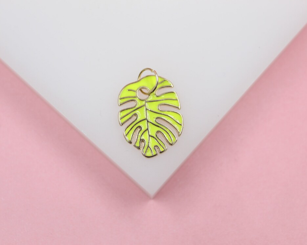 A neon green enameled monstera leaf charm rests on a white and pink display surface.