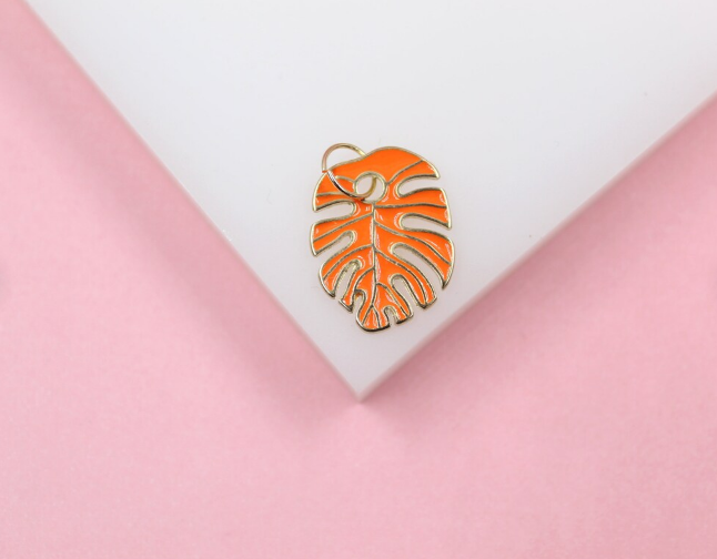 A vibrant orange monstera leaf pendant with gold trim rests on a white display against a soft pink background.