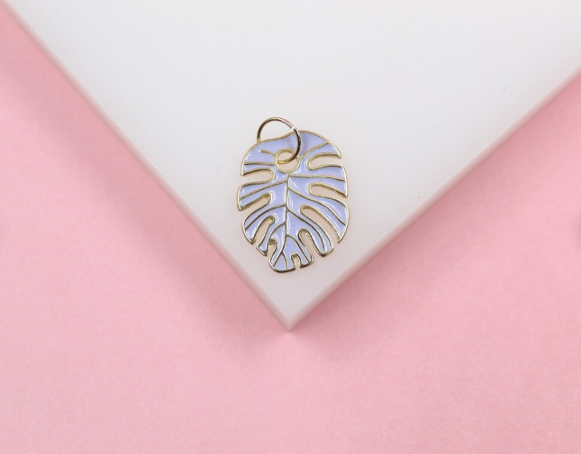 A light purple enamel monstera leaf charm with gold trim rests on a white display against a pink background.