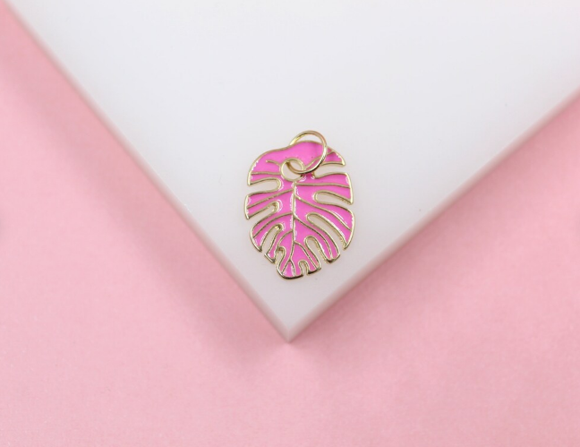 A gold-trimmed monstera leaf charm enameled in vibrant pink rests on a white display surface against a pink background.