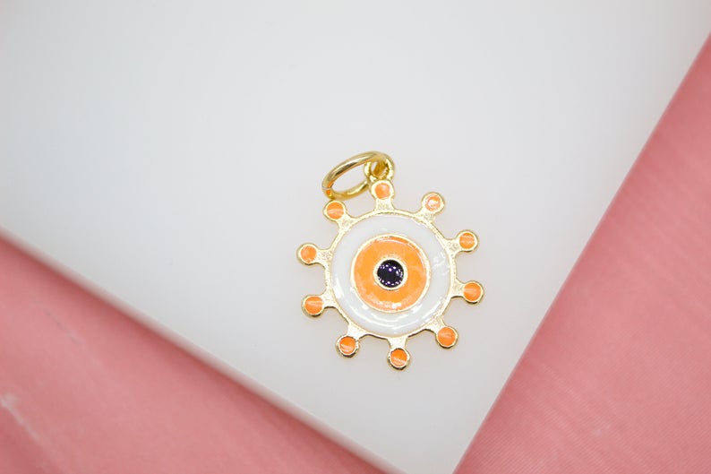 A gold-plated evil eye pendant features orange and white enamel with small rays radiating from the outer edge.