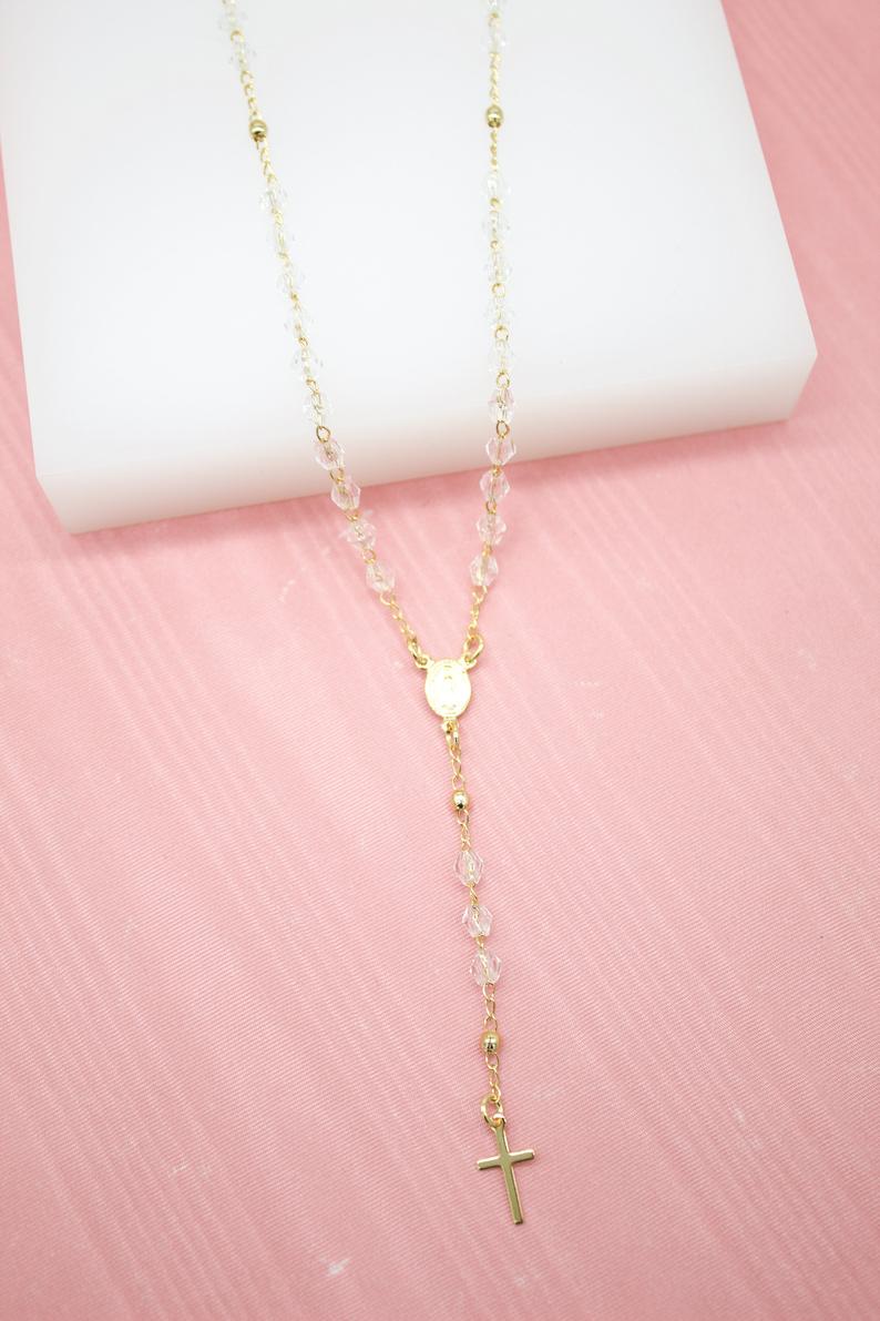 A delicate gold rosary necklace featuring clear crystal beads rests on a pink and white display surface.