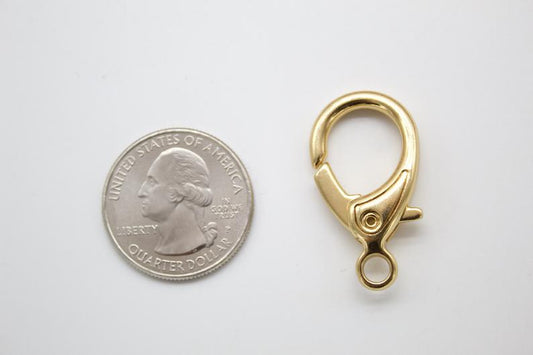 A shiny gold lobster clasp sits next to a US quarter coin for scale on a plain white background.