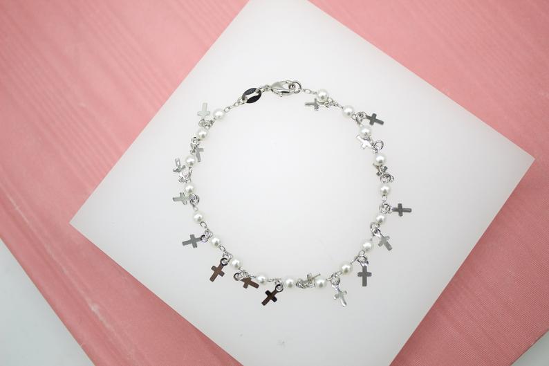 A delicate silver charm bracelet featuring hanging cross charms and small white pearls rests on a white display surface.
