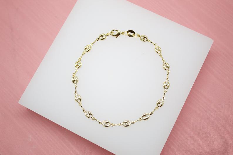 A delicate gold chain bracelet rests neatly on a white display pad against a soft pink background.