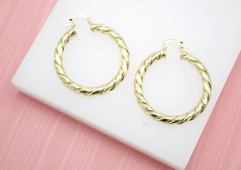 A pair of textured gold hoop earrings rests neatly on a white display stand.