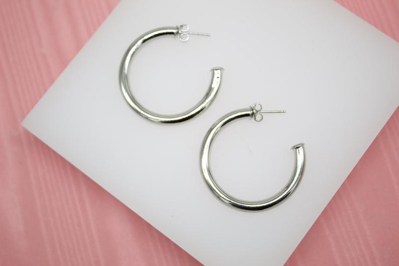 A pair of thick silver hoop earrings rests on a white display stand against a pink background.