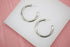 A pair of thick silver hoop earrings rests on a white display stand against a pink background.