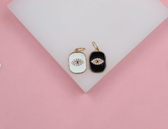 Two gold evil eye charms with white and black enamel sit on a white display against a pink background.