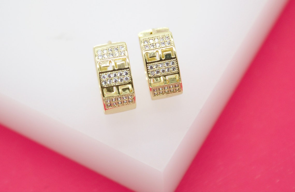 A pair of gleaming gold hoop earrings with intricate geometric cutouts and sparkling pave stones rest neatly on a white display against a vibrant pink backdrop.