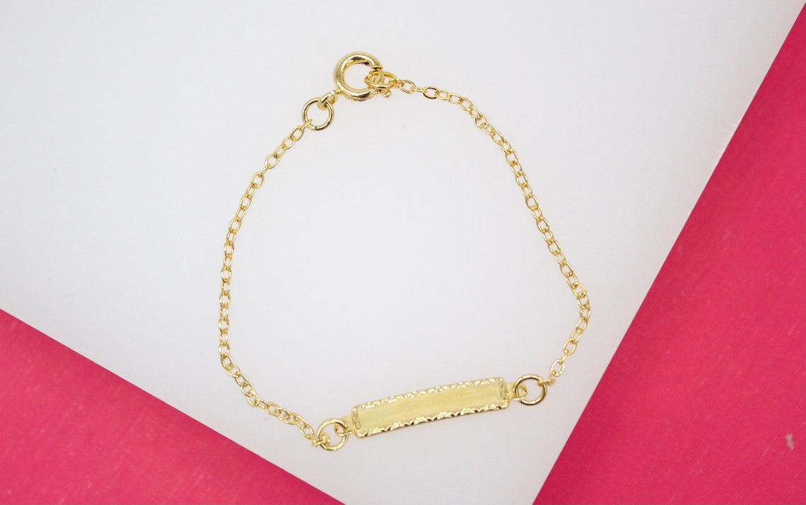 A delicate gold chain bracelet features an engraved ID bar resting against a white and pink background.