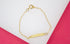 A delicate gold chain bracelet features an engraved ID bar resting against a white and pink background.