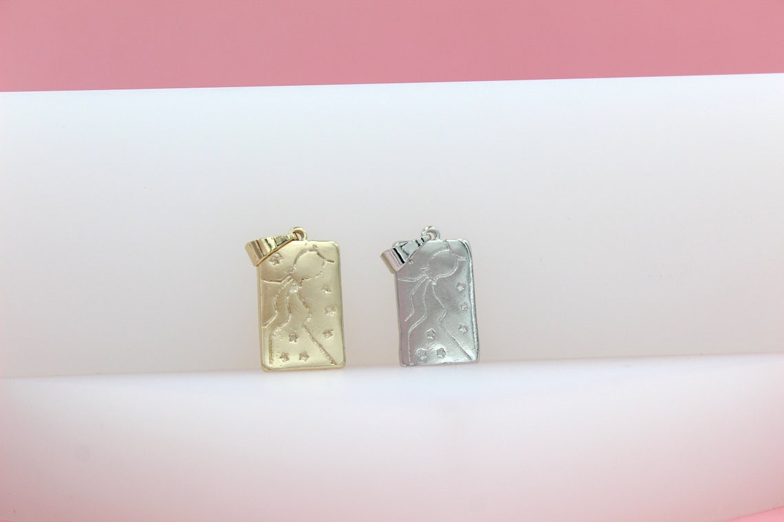 Two rectangular metal charms featuring engraved celestial figures rest side by side on a curved white display against a pink backdrop.