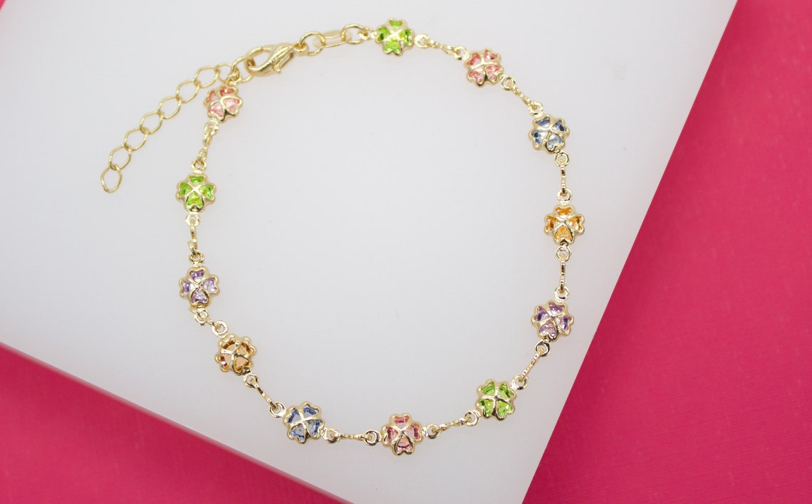 A delicate gold chain bracelet features colorful crystal four-leaf clover charms arranged against a white and pink background.
