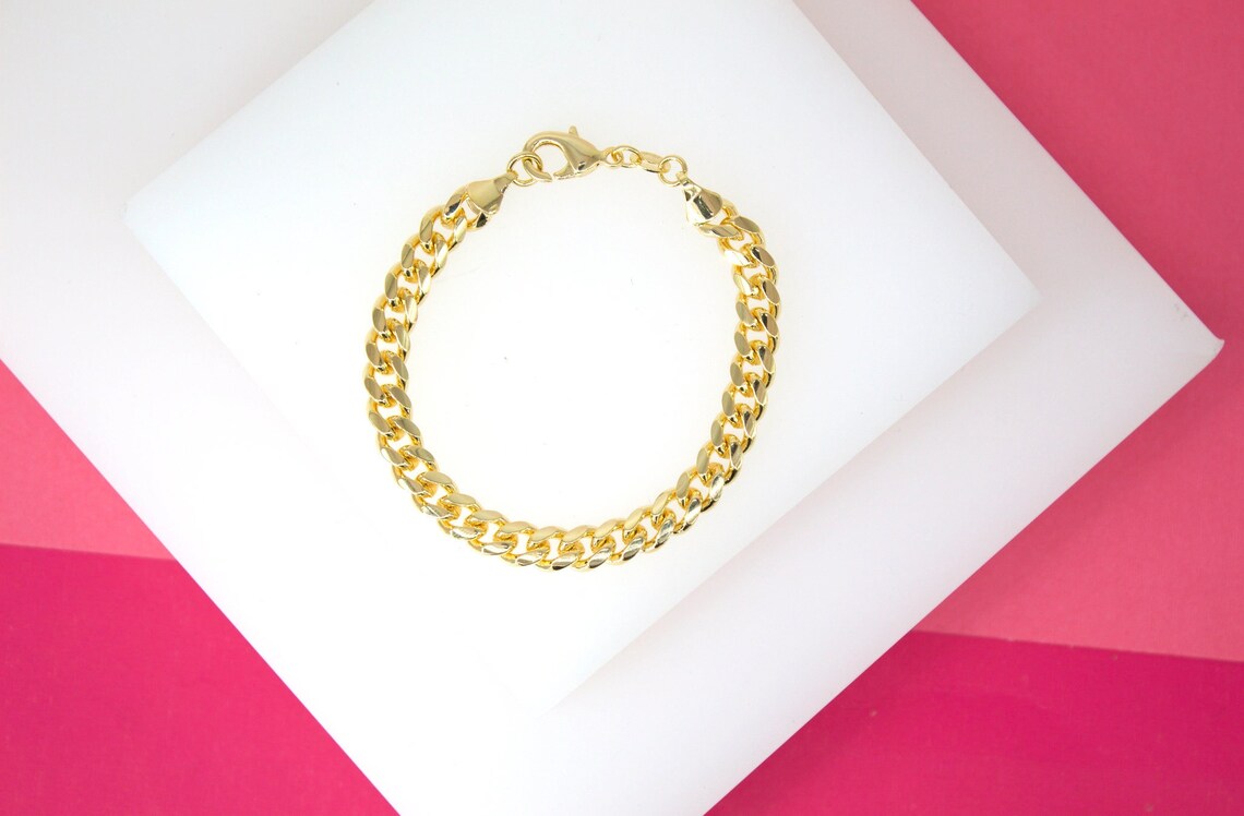 A polished gold curb chain bracelet rests elegantly on a white display block against a vibrant pink background.
