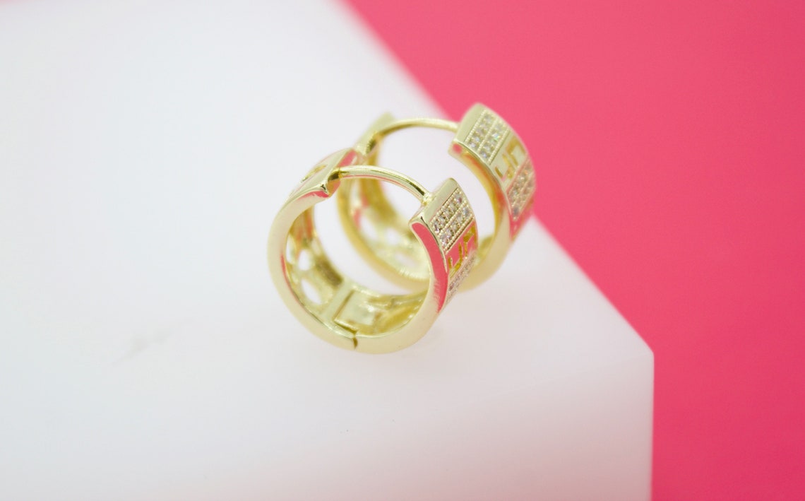A pair of sparkling gold hoop earrings resting on a white display block against a pink background.