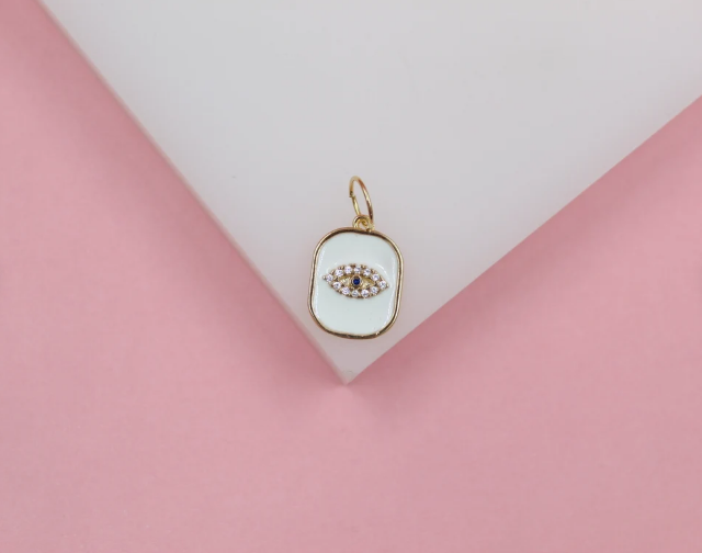 A gold and white enamel evil eye charm rests on a white geometric display against a soft pink background.