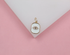 A gold and white enamel evil eye charm rests on a white geometric display against a soft pink background.