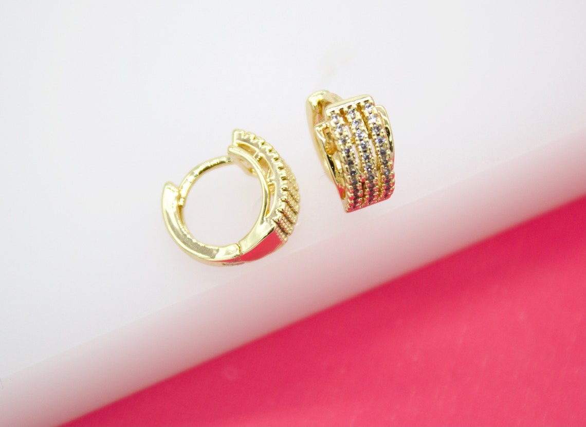 A pair of gold huggie hoop earrings featuring sparkling pave-set stones rest on a pink and white display surface.