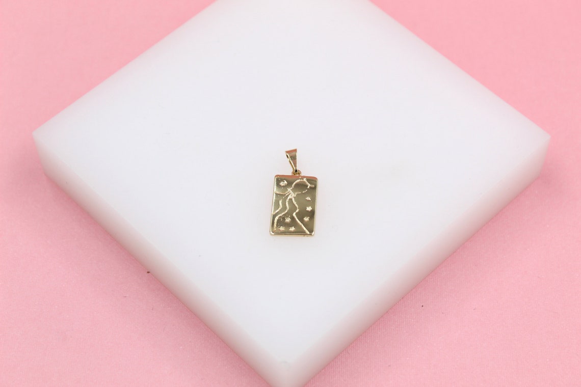 A gold rectangular pendant featuring an engraved celestial constellation rests on a white block against a soft pink background.