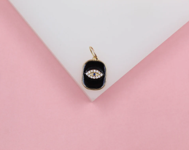A gold evil eye charm featuring sparkling cubic zirconia stones and black enamel rests on a white display surface against a pink background.