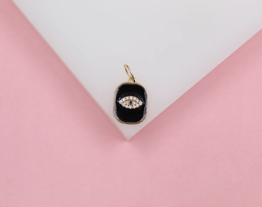 A gold evil eye charm featuring sparkling cubic zirconia stones and black enamel rests on a white display surface against a pink background.