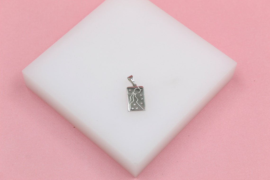 A silver rectangular Aquarius zodiac pendant rests on a white block against a pink background.
