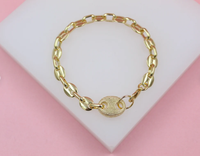 A gold-plated chain bracelet featuring textured oval links rests on a white display surface against a pink background.